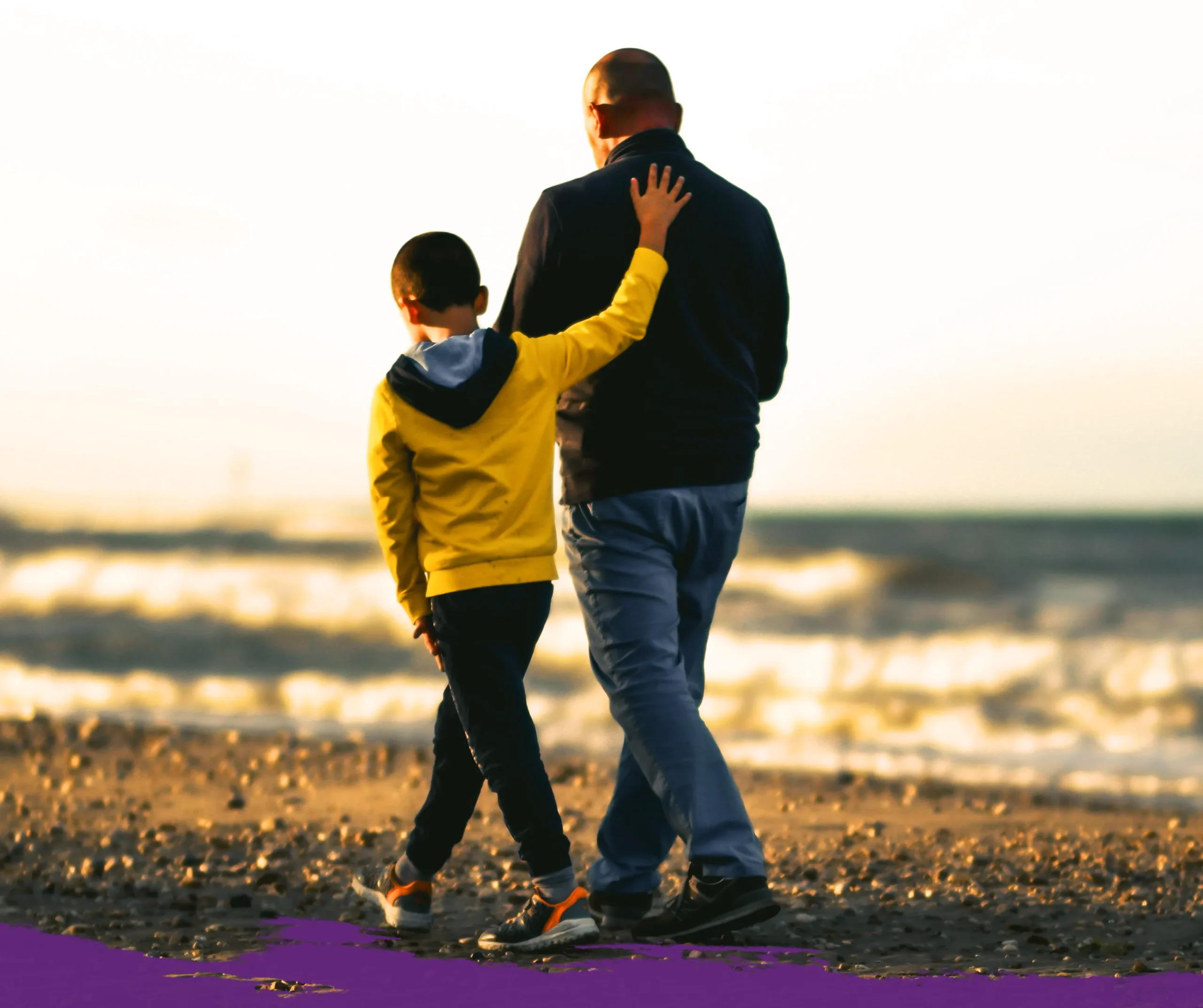 Father and small son walking along the shoreline away from the camera. Son has his hand on his father's back.
